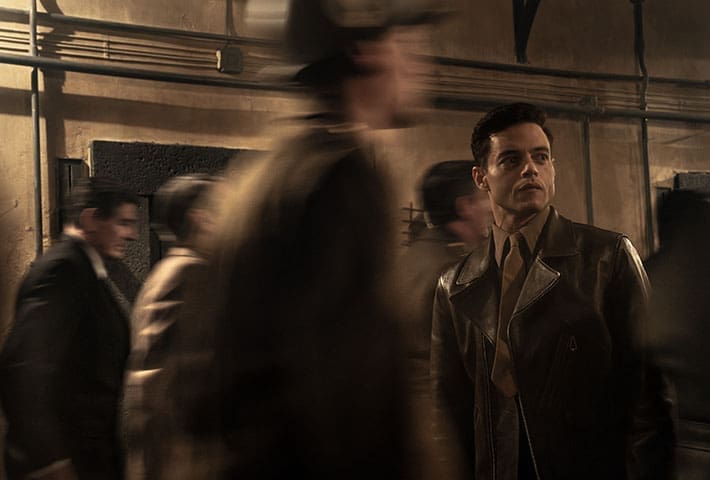 Rami Malek as psychiatrist Douglas Kelley in the film Nuremberg, he wears a leather overcoat over a military green tie and shirt, a number of people walk past him blurred in the image.