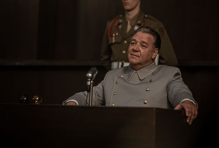 Russel Crowe as Hermann Göring in the film Nuremberg, sitting on the stand behind a microphone, there's a guard stood behind him in military uniform.
