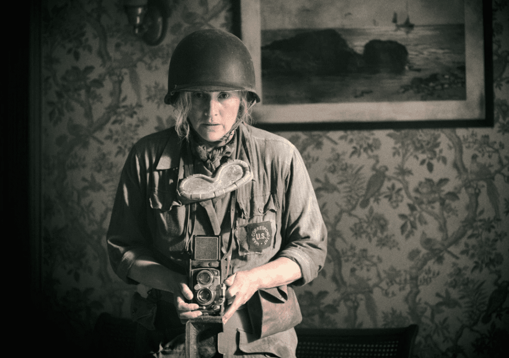 A woman in worn army uniform and a green rounded helmet stands holding a camera hanging around her neck