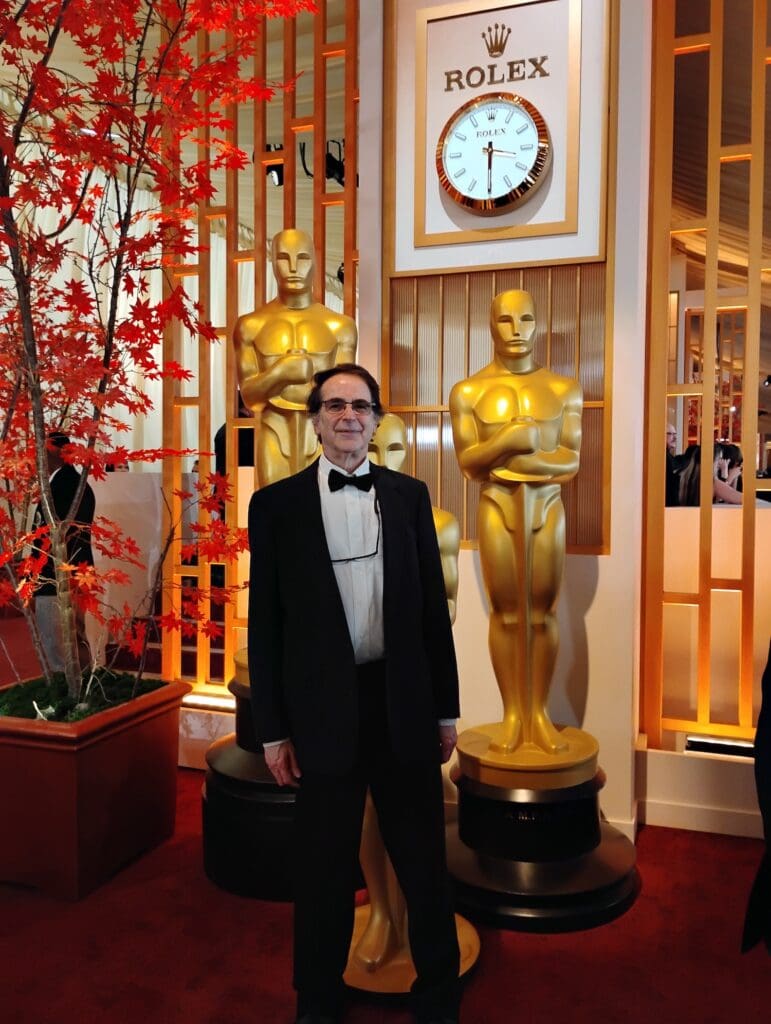 A man in a tuxedo stood on a red carpet in front of three Oscar statuettes taller than him