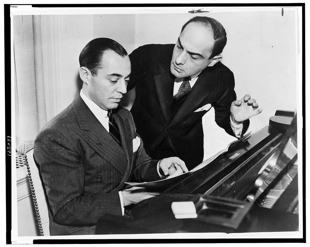 A man sat at a piano reading lyrics, there's another man stood next to him standing up gesticulating with his hand.
