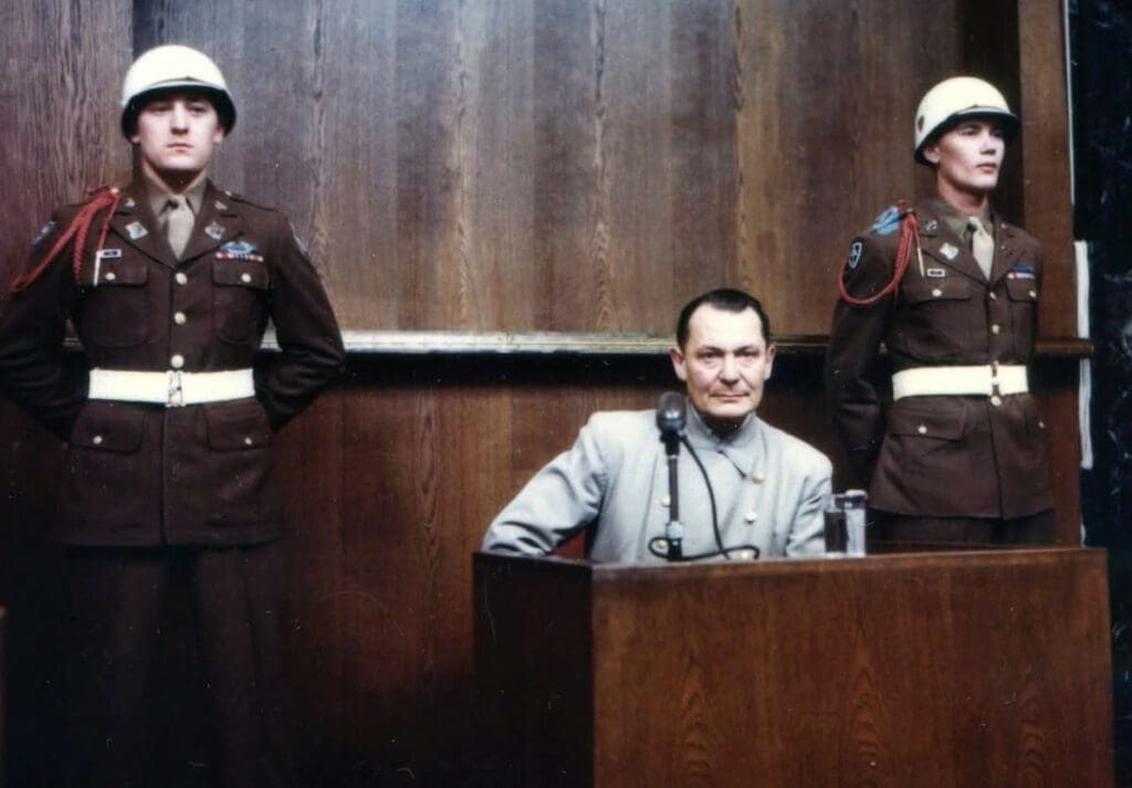 A real picture of Hermann Göring during the Nuremberg trial, similar to the picture of Russel Crowe next to it he sits at the stand behind the microphone in a blue shirt, with two uniformed guards stood either side of him.