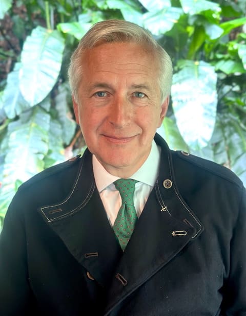 A man in a blue overcoat with a green tie looks directly at the camera smiling