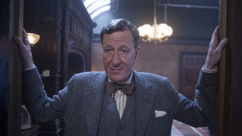 a man in a three  piece grey suit looks down the camera opening a door with both hands
