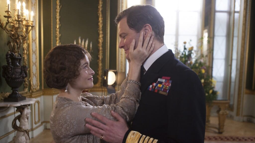 A woman holds the face of a man in a black military uniform, they're in a lavishly decorated room in a royal palace
